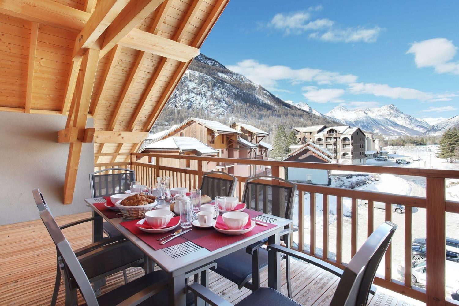 Przytulny balkon z drewnianym zadaszeniem i stołem nakrytym do śniadania, z widokiem na ośnieżone Alpy i miasteczko Serre Chevalier we Francji – komfortowe wakacje z biurem podróży promoclub.pl