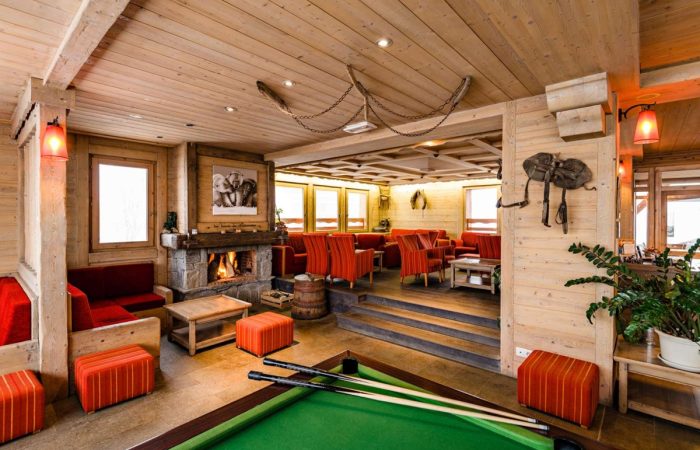 Stylowy alpejski lounge z kominkiem – ciepłe drewno, kamienne detale i przytulne fotele w odcieniach czerwieni tworzą idealne miejsce na relaks po dniu spędzonym na stoku