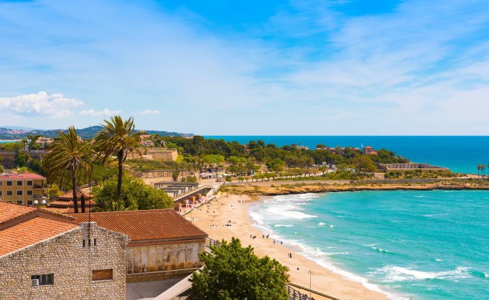 Słoneczna plaża w Hiszpanii z turkusowym morzem, palmami i historycznymi zabudowaniami w tle, idealne miejsce na wakacje i relaks nad Morzem Śródziemnym, promoclub.pl