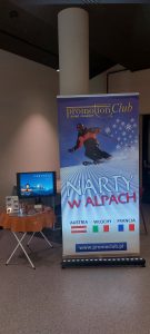 Stoisko biura podróży Promoclub z podświetlonym banerem reklamowym „Narty w Alpach”, przedstawiającym narciarza na ośnieżonym stoku. Obok znajduje się stolik z monitorem wyświetlającym zdjęcie gór i materiałami informacyjnymi o wyjazdach do Austrii, Włoch i Francji – promoclub.pl