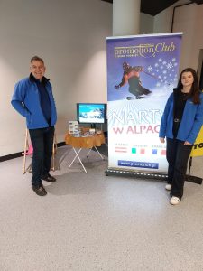 Dwoje pracowników biura podróży Promoclub stoi przy stoisku promocyjnym z banerem „Narty w Alpach”. Na banerze widać narciarza na stoku oraz nazwy krajów: Austria, Włochy, Francja. Obok znajduje się stolik z monitorem prezentującym ofertę wyjazdów zimowych – promoclub.pl